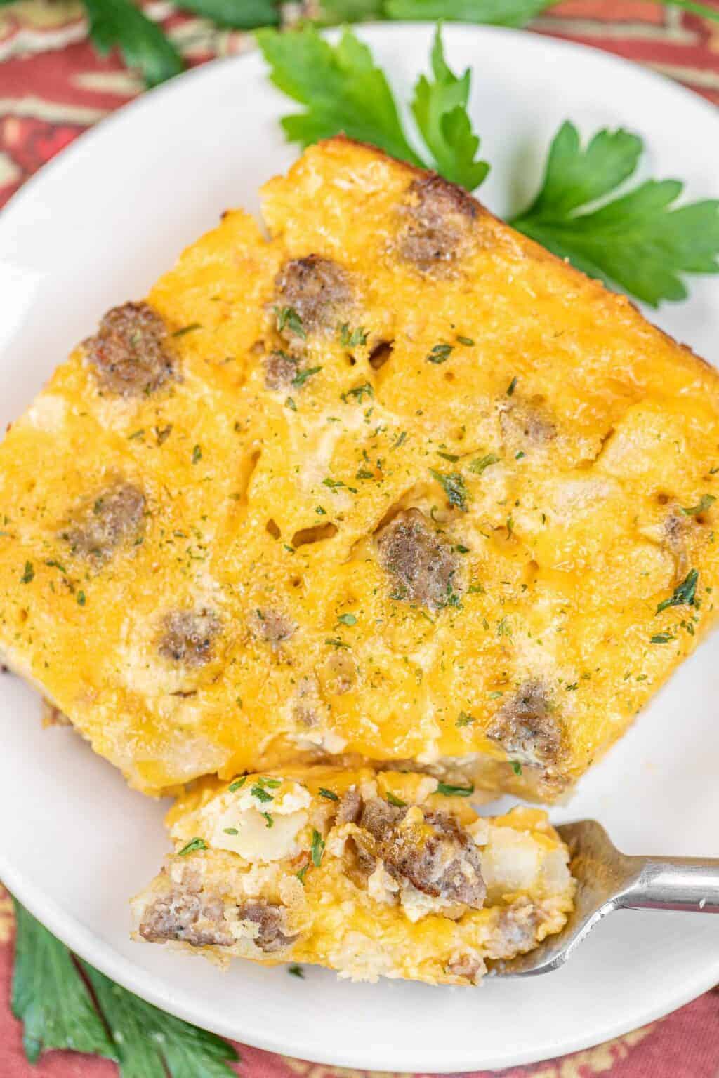 A close-up of a cheesy breakfast casserole with sausage and eggs, served on a white plate and garnished with fresh parsley. A fork holds a bite-sized piece of the casserole in the foreground.