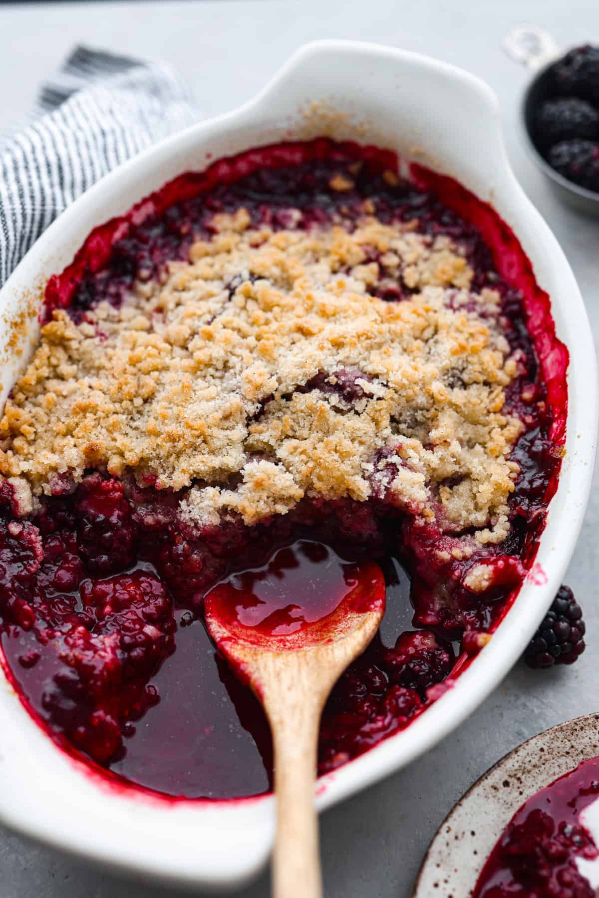 A white baking dish filled with blackberry crumble, topped with golden crumbly streusel and juicy blackberry filling, with a wooden spoon scooping out a portion. A striped towel and fresh blackberries are nearby.