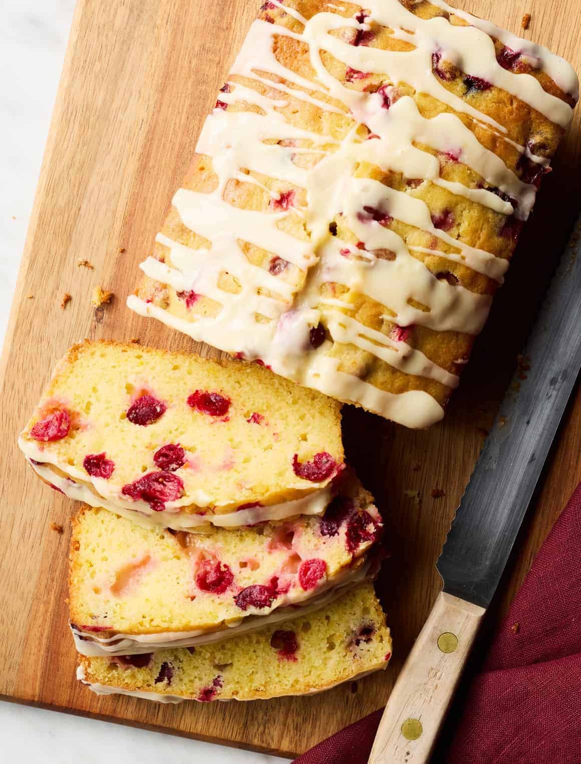 A cranberry loaf cake drizzled with white icing sits on a wooden cutting board. Two slices are cut, showing cranberries inside. A serrated knife with a wooden handle lies next to the cake.