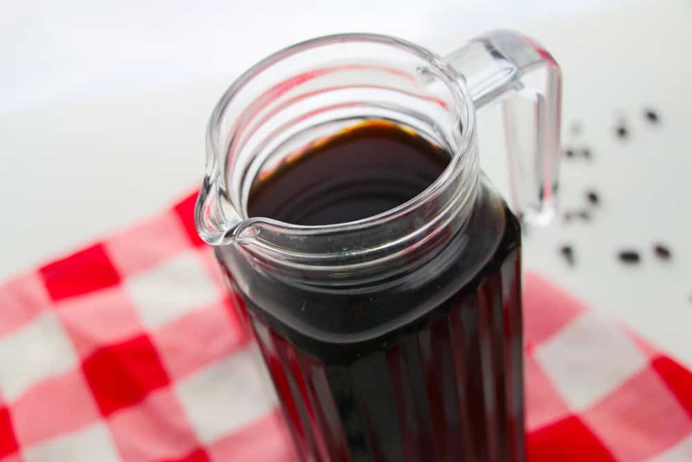 A clear glass pitcher filled with dark liquid, possibly a cold brew recipe or iced tea, sits on a red and white checkered cloth with a few black seeds scattered nearby.