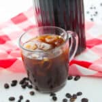 A glass mug of iced coffee, inspired by a cold brew recipe, sits on a white surface with scattered coffee beans, in front of a bottle of coffee concentrate and a red and white checkered cloth.