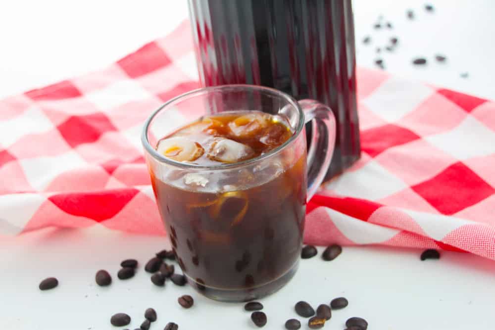 A glass mug of iced coffee, inspired by a cold brew recipe, sits on a white surface with scattered coffee beans, in front of a bottle of coffee concentrate and a red and white checkered cloth.