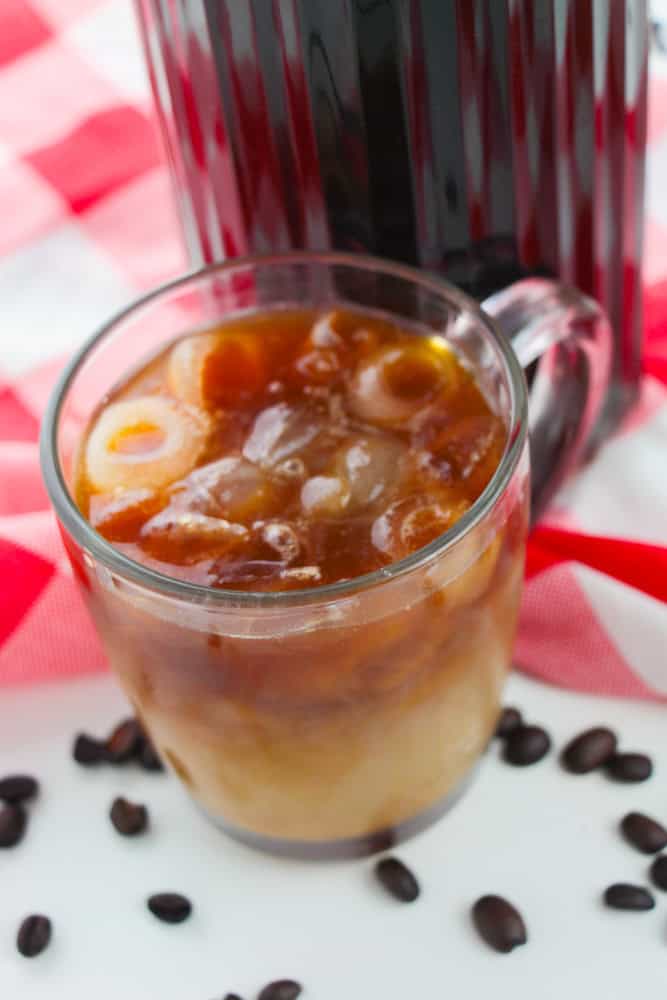 A glass of iced coffee, perfect for a cold brew recipe, sits on a white surface with visible ice cubes and scattered coffee beans. Behind it rests a large container of dark coffee and a red and white checkered cloth.