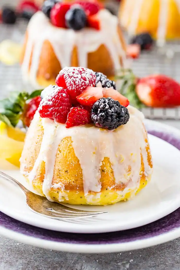 A mini bundt cake drizzled with white icing, topped with fresh berries and a dusting of powdered sugar, sits on a white plate with a fork. A sliced strawberry and another cake appear in the background.