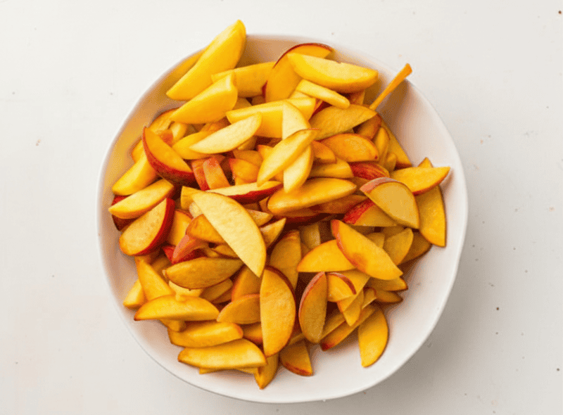 A white bowl filled with thinly sliced apple pieces, some with red skin, on a plain white surface—perfect for adding to your favorite peach cobbler recipe.