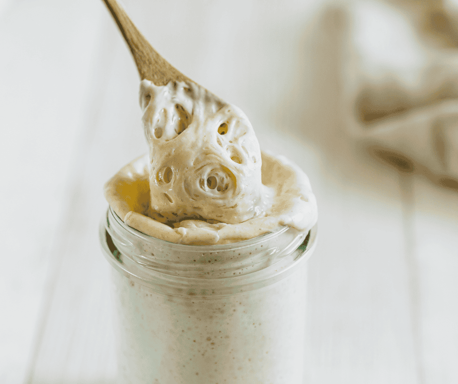 A wooden spoon lifts thick, creamy mayonnaise from a glass jar, with some of the spread spilling over the rim—perfect for slathering on freshly baked sourdough biscuits. The background is light and softly focused.