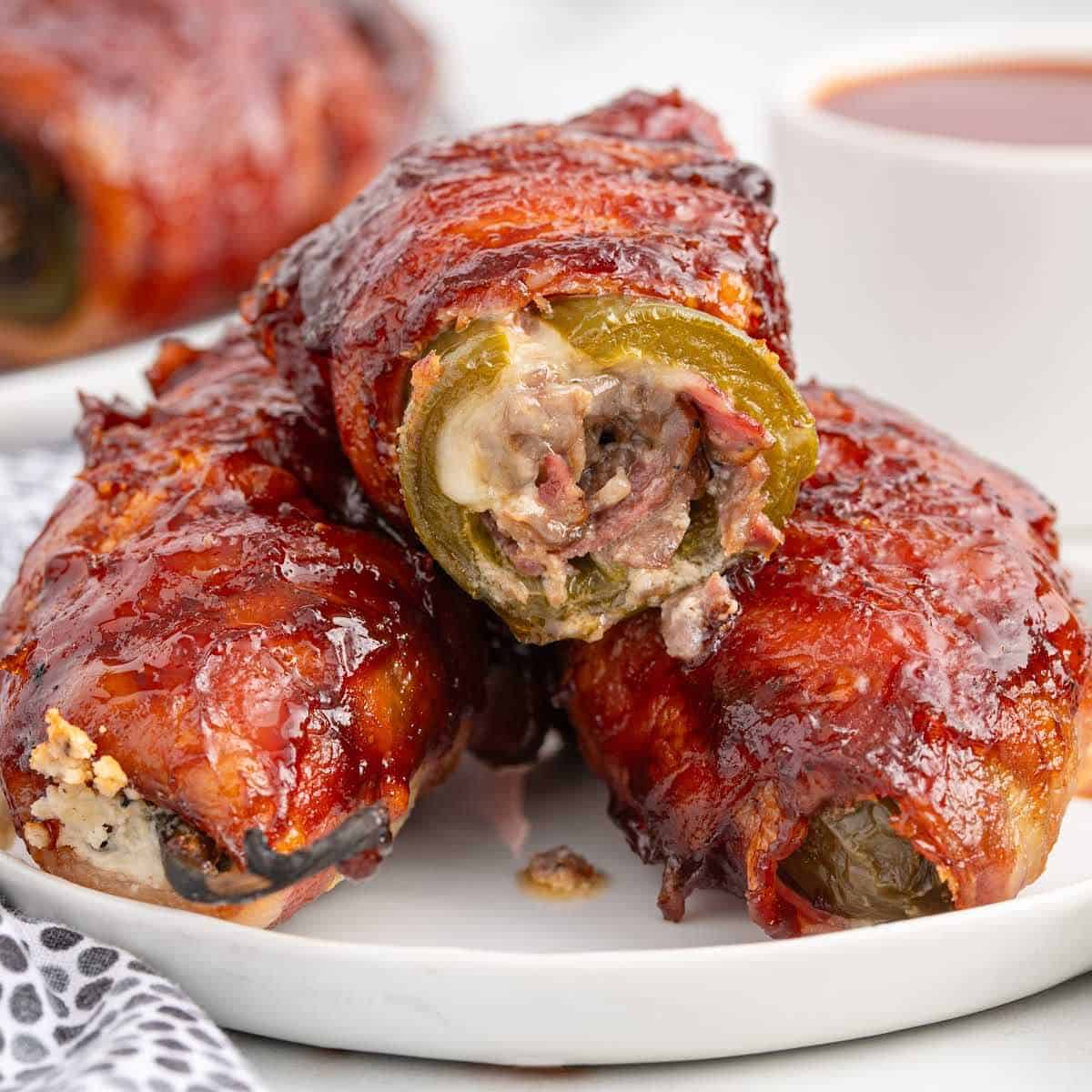 Three bacon-wrapped, cheese and meat-stuffed jalapeños are stacked on a white plate, coated in a glossy barbecue sauce, with a cup of dipping sauce blurred in the background.