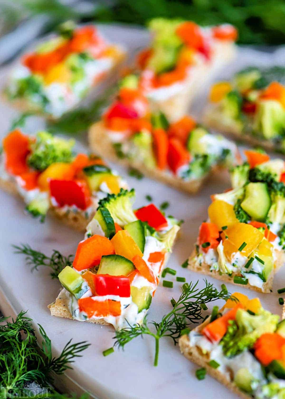 Rectangular crackers topped with a creamy spread and colorful chopped vegetables, including broccoli, red bell pepper, yellow bell pepper, and carrot, displayed on a white platter with fresh herbs.
