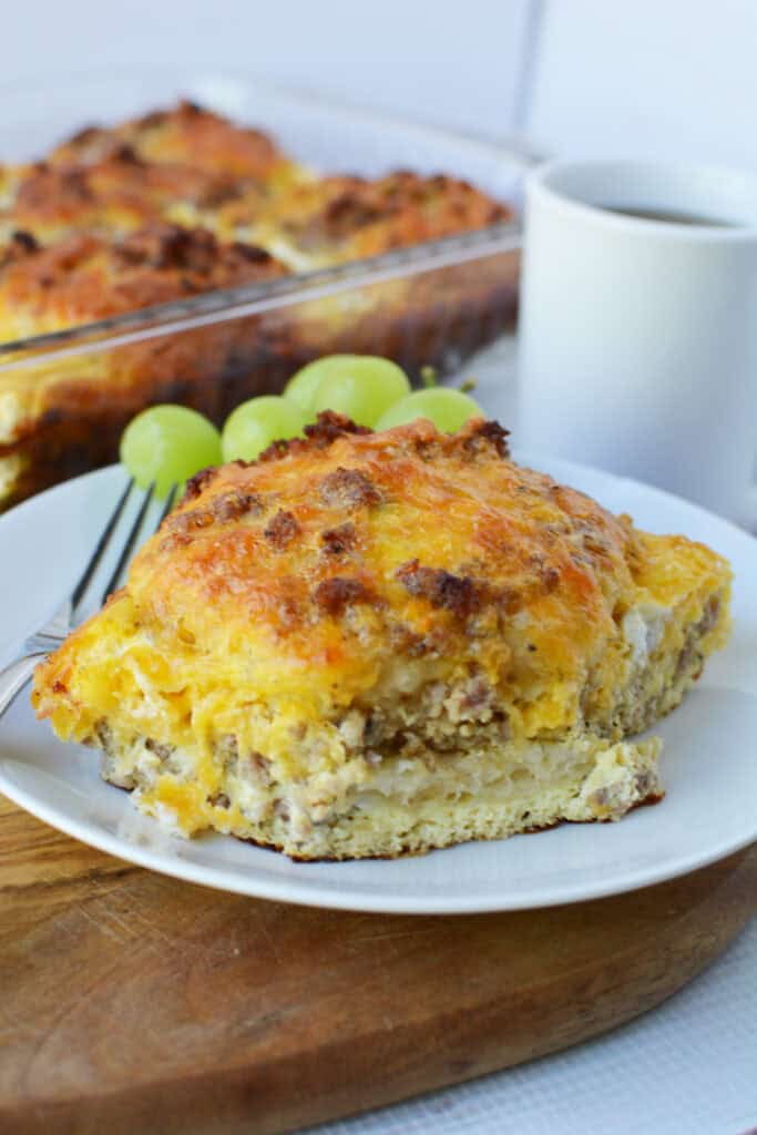 A slice of cheesy Biscuit Breakfast Casserole with sausage sits on a white plate, served with green grapes. A fork, a white mug, and a casserole dish are in the background.