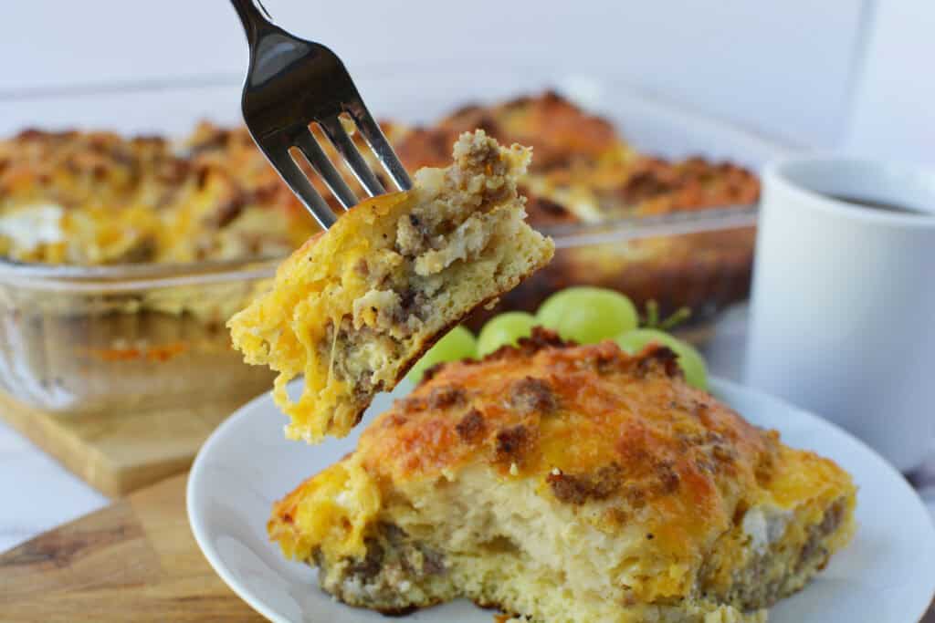 A fork holds a bite of cheesy Biscuit Breakfast Casserole with sausage and eggs above a plate and glass baking dish, with grapes and a cup in the background.