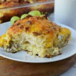 A plate with a serving of Biscuit Breakfast Casserole, featuring eggs, sausage, and melted cheese, sits on a wooden board. Green grapes and a white mug are in the background.