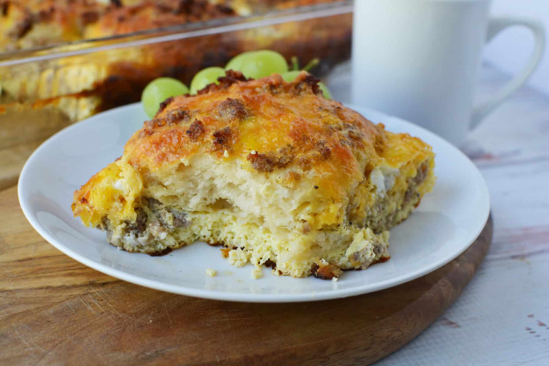 A plate with a serving of Biscuit Breakfast Casserole, featuring eggs, sausage, and melted cheese, sits on a wooden board. Green grapes and a white mug are in the background.