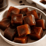 A white bowl filled with dark brown coffee ice cubes sits on a tan surface, with an ice cube tray and coffee beans visible in the background.