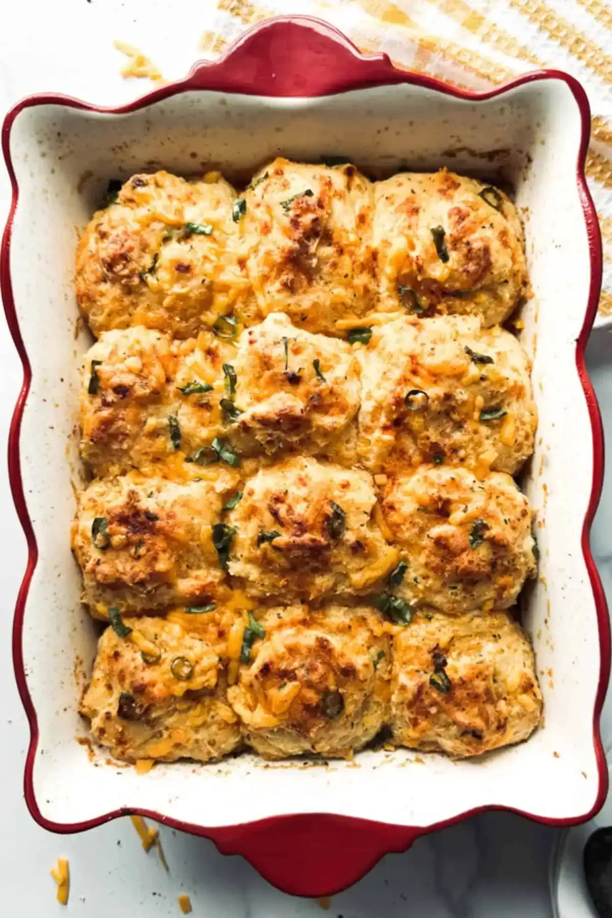 A red ceramic baking dish filled with twelve golden-brown, cheesy biscuits garnished with herbs, set on a white surface next to a yellow-striped towel.