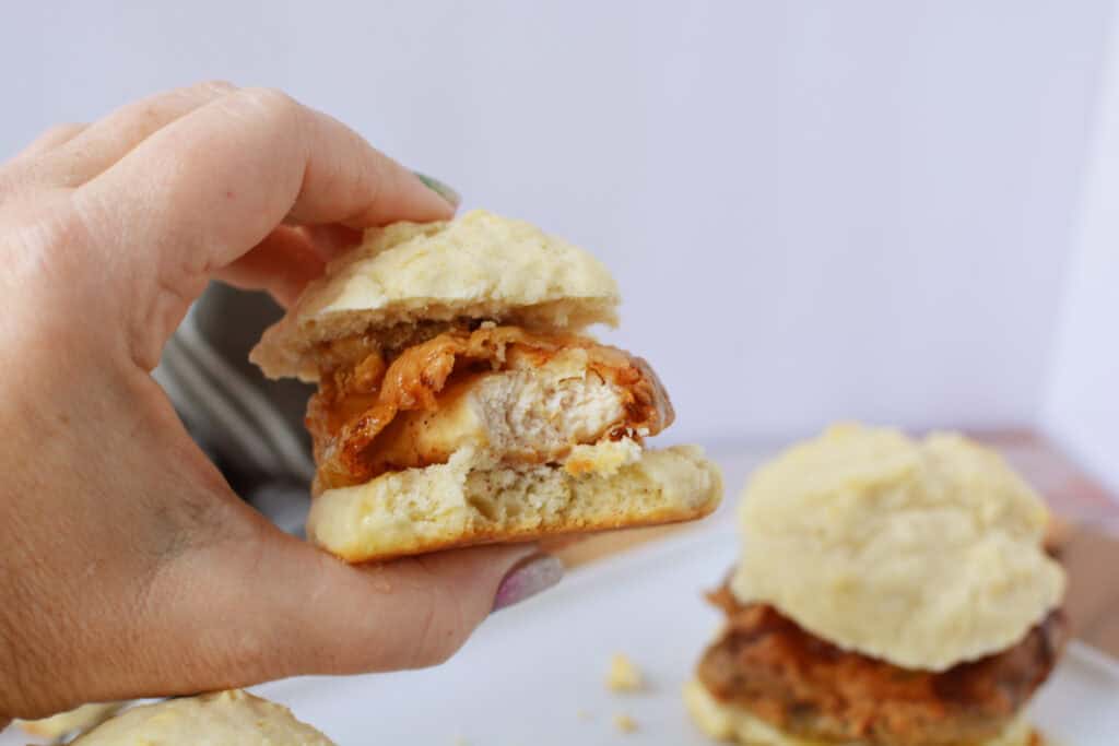 A hand holding a tempting Chicken Biscuit Sandwich filled with fried chicken and cheese; more biscuit sandwiches are visible in the background on a white surface, perfect for trying your next Chicken Biscuit Sandwich Recipe.