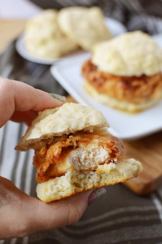 A hand holds a partially eaten chicken biscuit sandwich, inspired by a classic Chicken Biscuit Sandwich Recipe. In the background, two more sandwiches rest on a white plate atop a striped cloth.