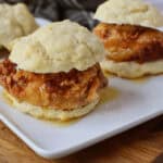 A close-up of fried chicken sandwiched between two halves of a flaky biscuit, drizzled with syrup, served on a white rectangular plate—this Chicken Biscuit Sandwich Recipe shines with extra biscuit sandwiches in the background.