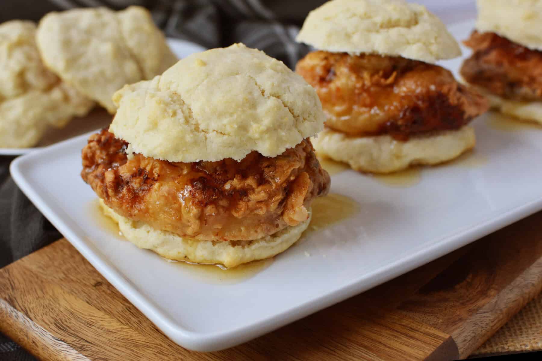 A close-up of fried chicken sandwiched between two halves of a flaky biscuit, drizzled with syrup, served on a white rectangular plate—this Chicken Biscuit Sandwich Recipe shines with extra biscuit sandwiches in the background.
