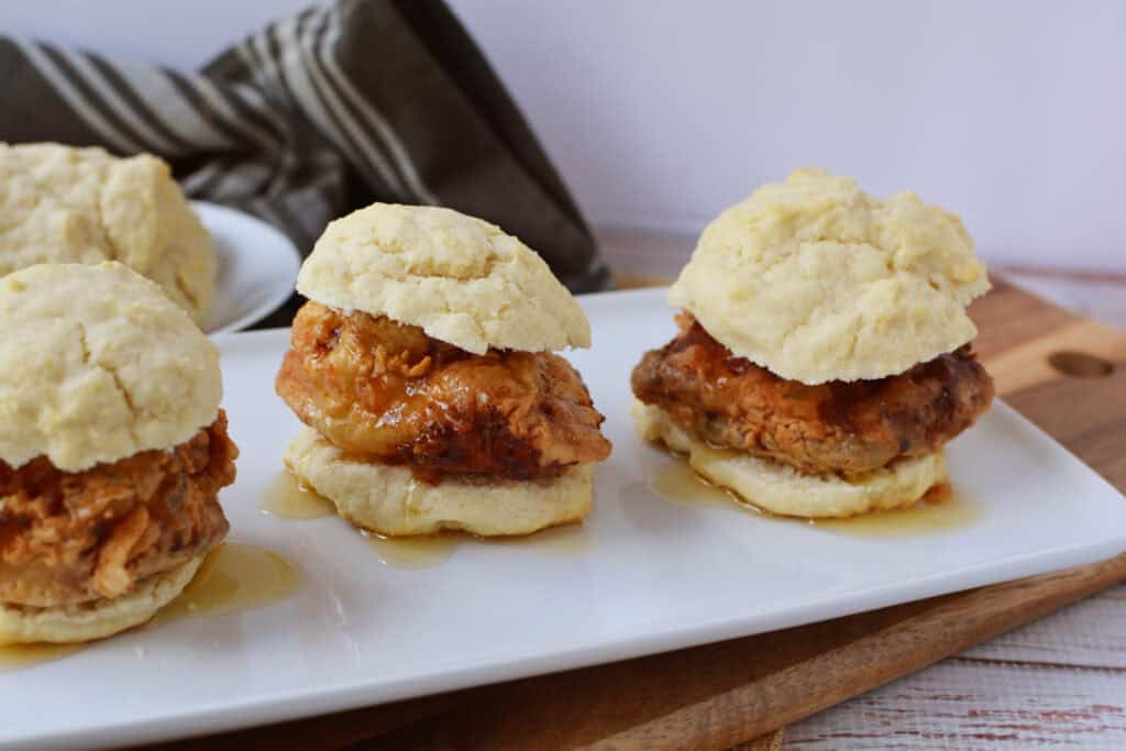 Three fried chicken biscuits with gravy are arranged on a white rectangular plate, illustrating a delicious Chicken Biscuit Sandwich Recipe, with a napkin and extra biscuits in the background on a wooden surface.