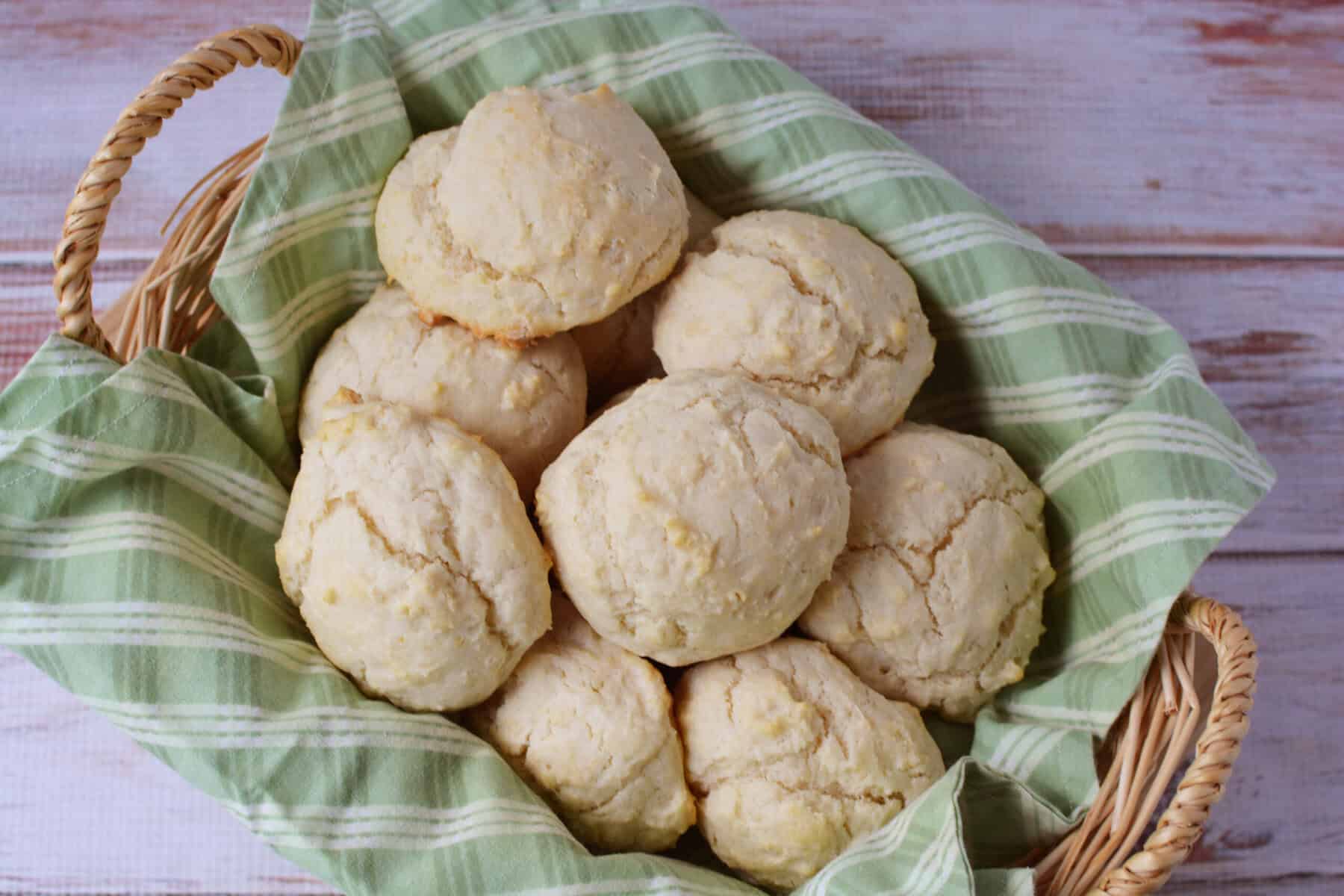 A basket lined with a green and white striped cloth, filled with round, golden-brown biscuits made from a classic Drop Biscuit Recipe, sits atop a light wooden surface.