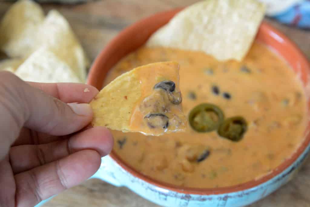 A hand holds a tortilla chip dipped in creamy cheese queso with black olives, above a bowl of the same dip garnished with sliced jalapeños.