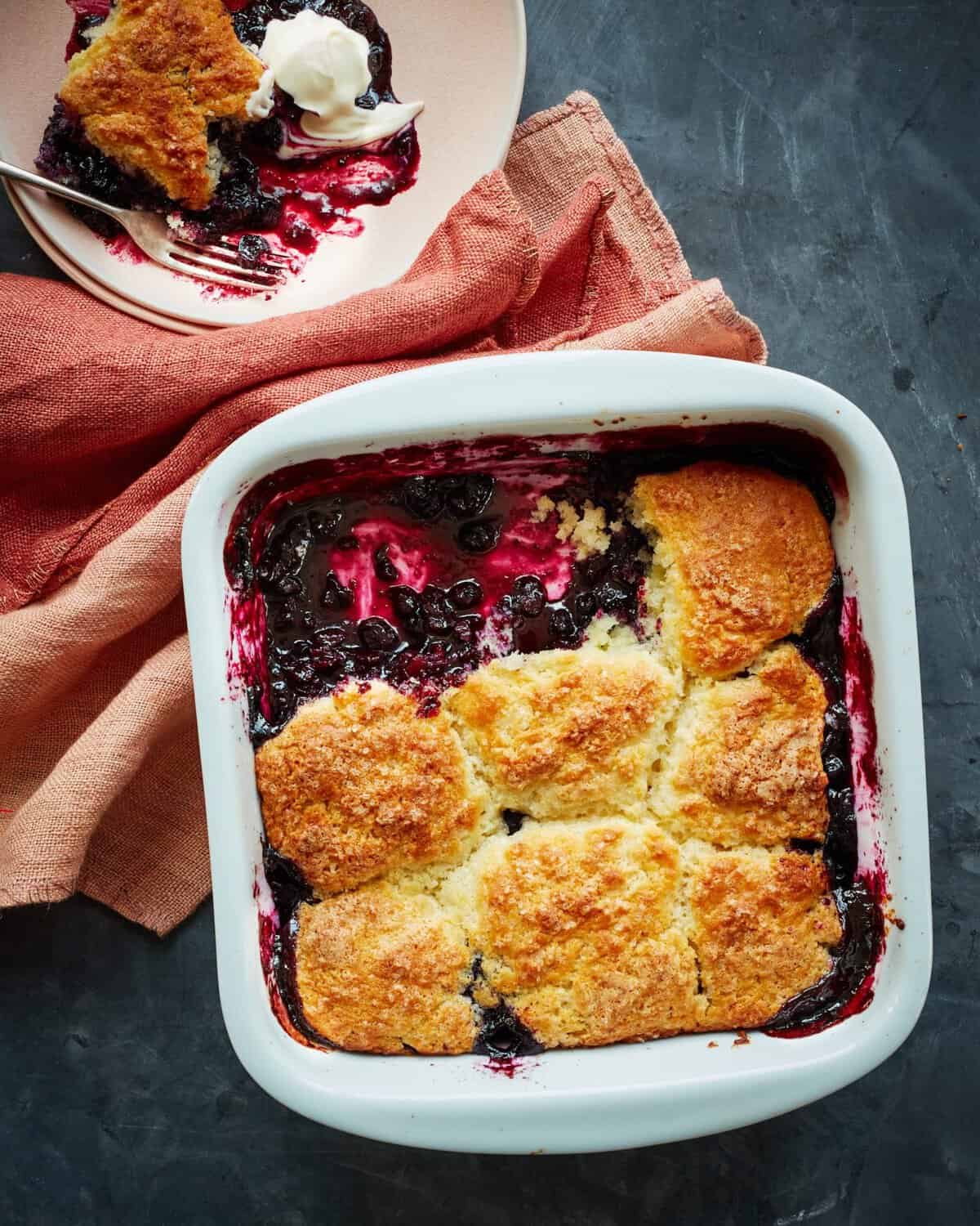 A baked blueberry cobbler in a white dish with golden biscuit topping, some served on a plate with a dollop of cream. The dish and plate rest on textured orange and pink cloths atop a dark surface.