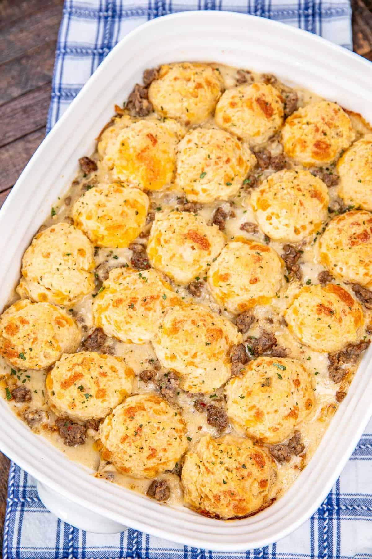 A white casserole dish filled with golden-brown biscuits on top of a creamy mixture with ground sausage, resting on a blue and white checkered cloth.