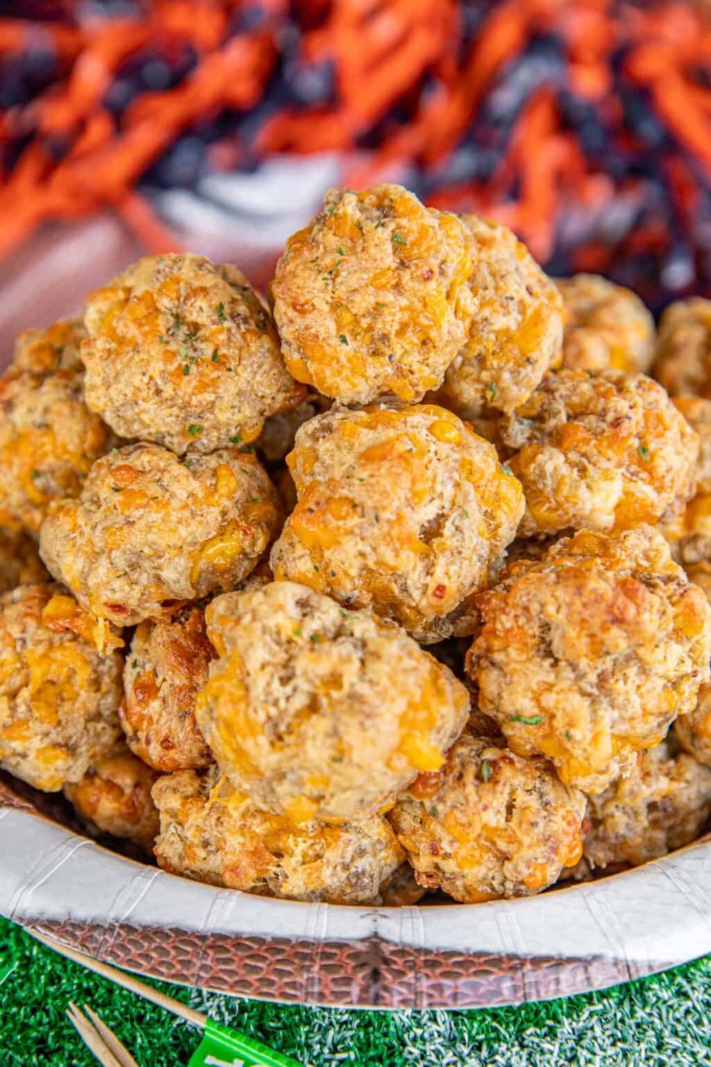 A close-up of a plate piled high with golden-brown sausage balls, showing bits of cheese and herbs, set on a sports-themed table with a blurred background.