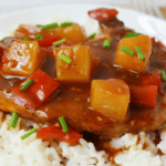 A plate of white rice topped with Hawaiian Pork Chops Slow Cooker style—tender meat in brown sauce, pineapple chunks, red bell pepper pieces, and a garnish of chopped green onions.