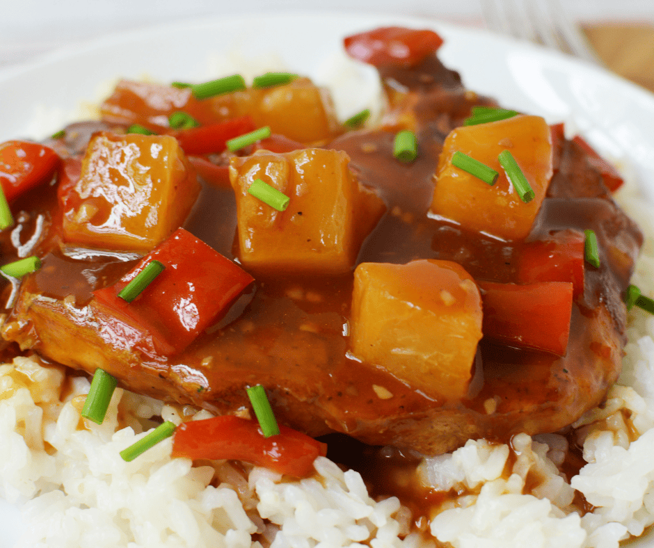 A plate of white rice topped with Hawaiian Pork Chops Slow Cooker style—tender meat in brown sauce, pineapple chunks, red bell pepper pieces, and a garnish of chopped green onions.
