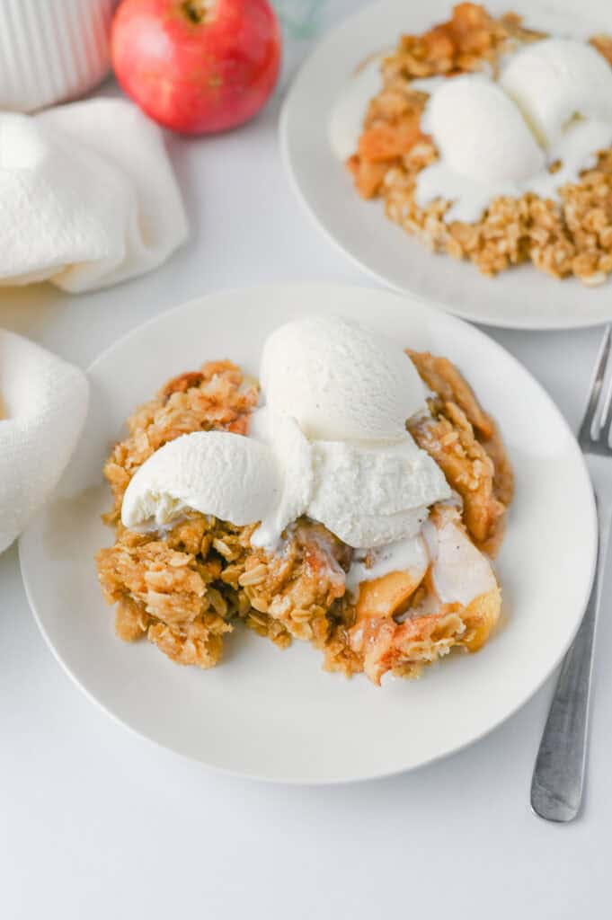 A white plate with a serving of Crock Pot Apple Crisp topped with three scoops of vanilla ice cream sits on a white table next to a fork. Another plate of apple crisp and a red apple are visible in the background.