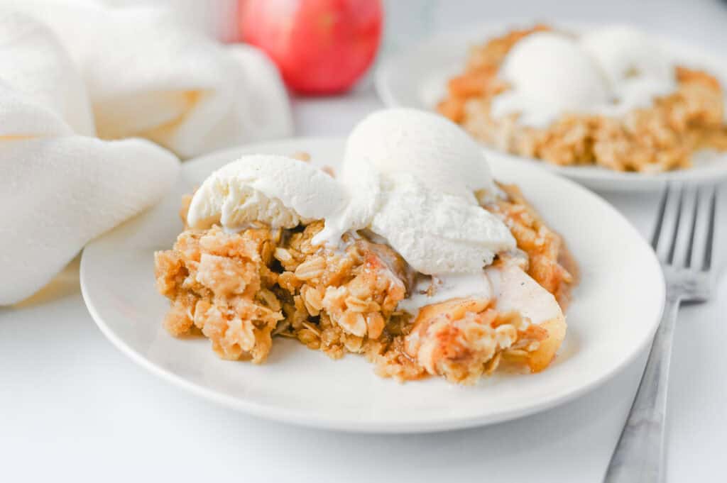 A white plate with Crock Pot Apple Crisp topped with three scoops of vanilla ice cream. Another plate with the same dessert sits in the background, along with a red apple and a fork on a white surface.