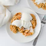 A plate of Crock Pot Apple Crisp topped with two scoops of vanilla ice cream sits next to a fork, with a white napkin nearby and another plate of dessert partially visible.