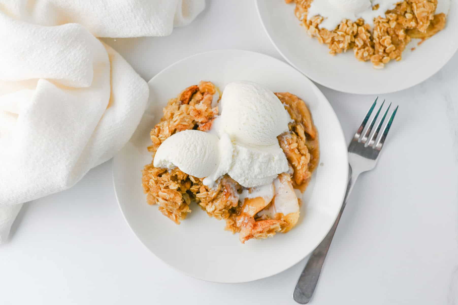 A plate of Crock Pot Apple Crisp topped with two scoops of vanilla ice cream sits next to a fork, with a white napkin nearby and another plate of dessert partially visible.