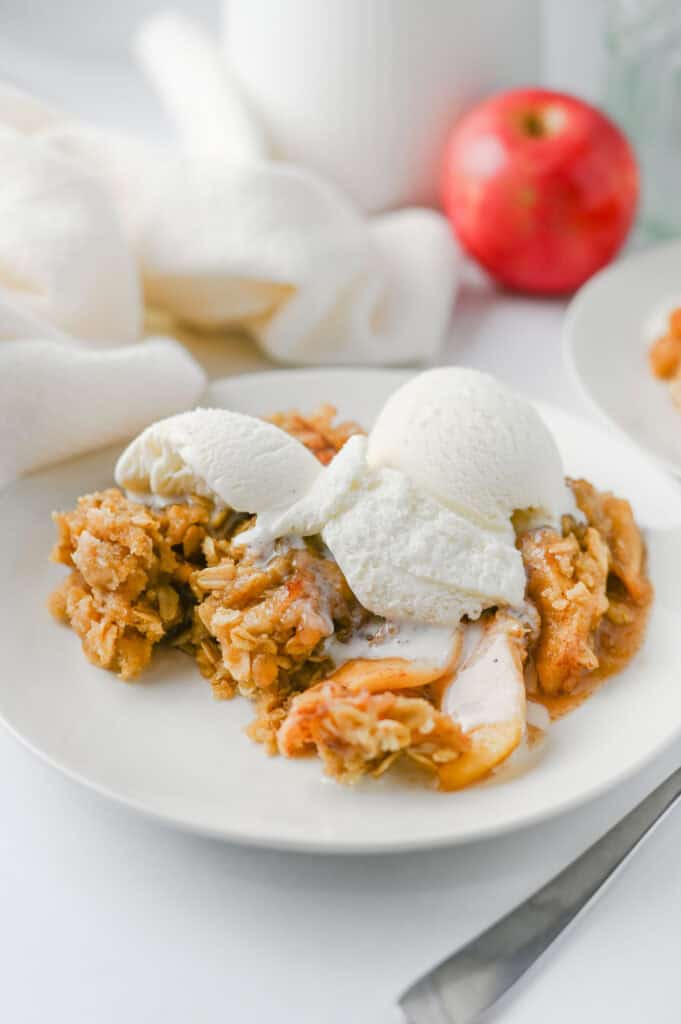A plate of Crock Pot Apple Crisp topped with two scoops of vanilla ice cream sits on a white table. A fresh red apple and a white cloth are in the background.