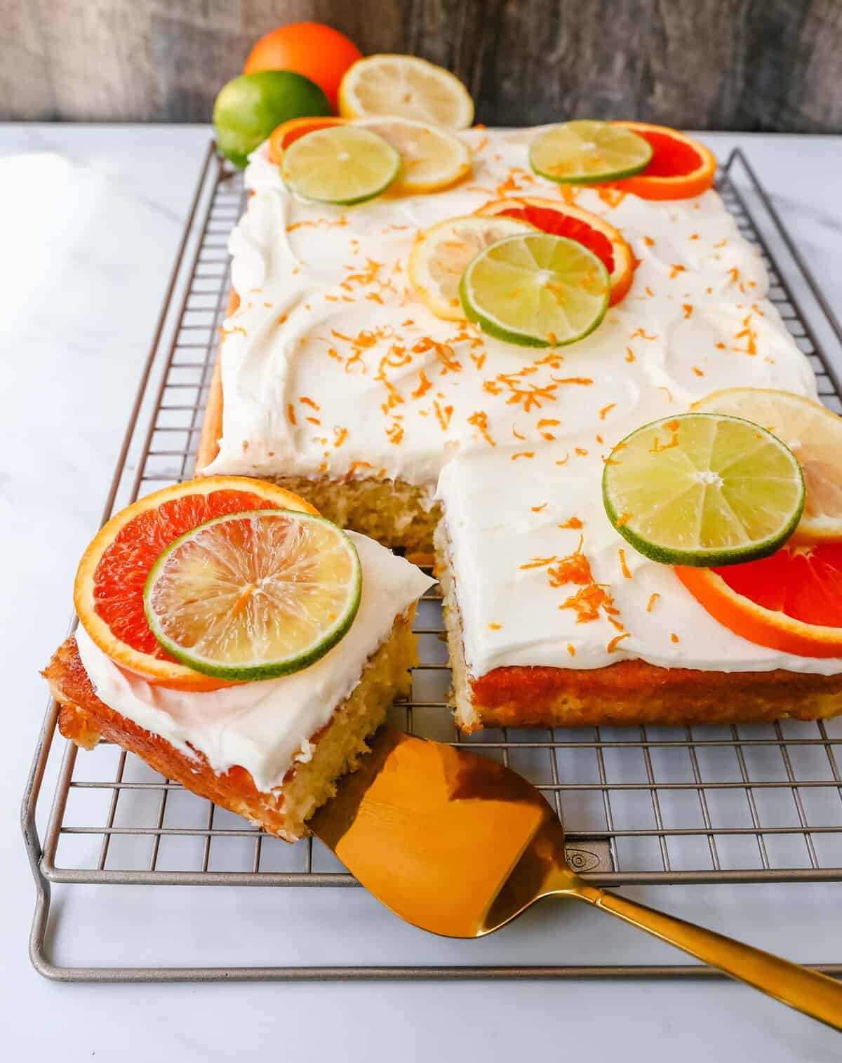 A frosted sheet cake on a cooling rack, topped with lime, lemon, and orange slices plus orange zest—perfect for summer birthday cakes. A square piece has been cut and is being lifted with a gold-colored cake server.
