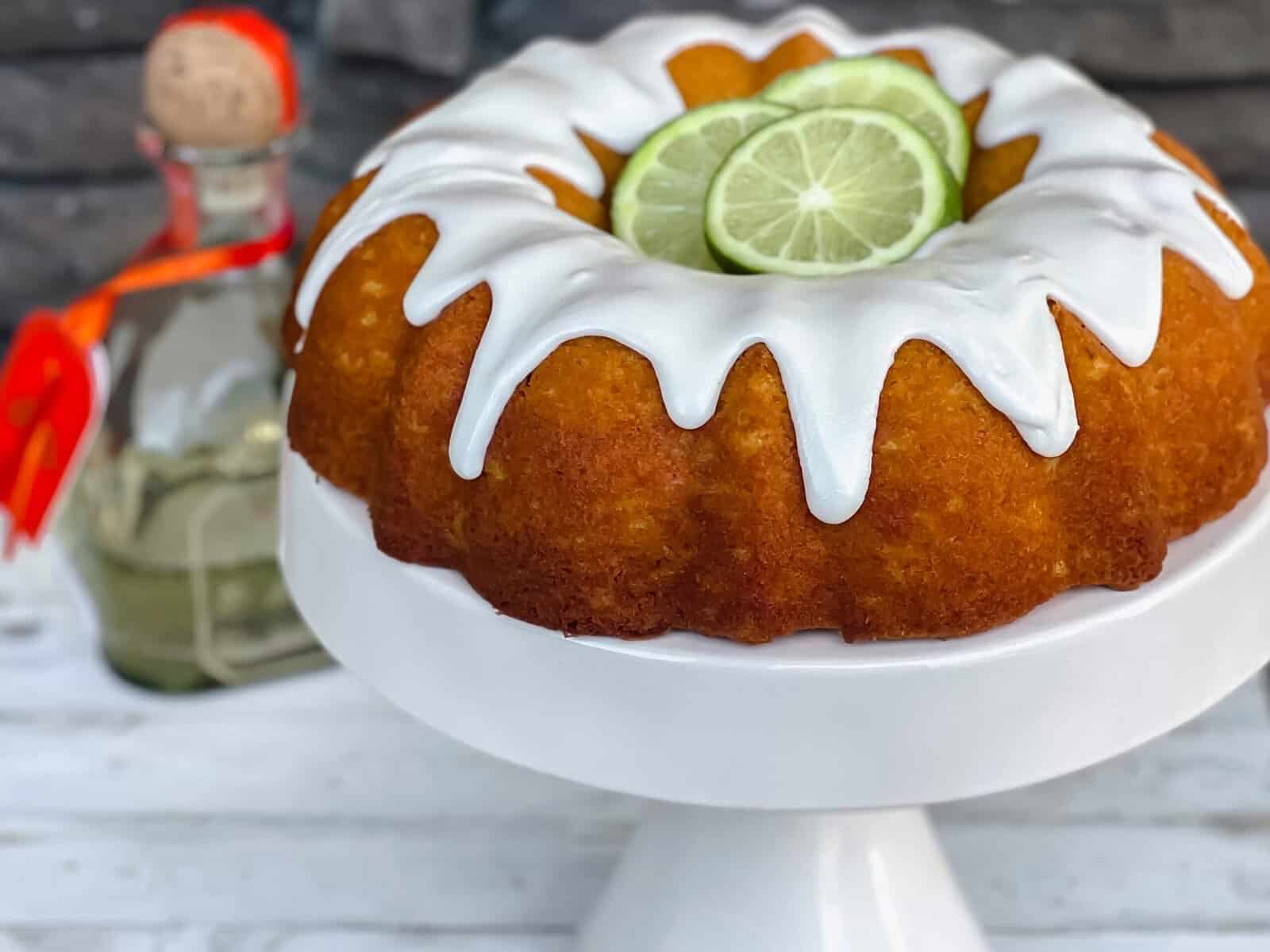 A bundt cake with white icing drips, topped with three lime slices, sits on a white cake stand. In the background, there’s a bottle with a cork and red ribbon, all set against a gray stone wall.