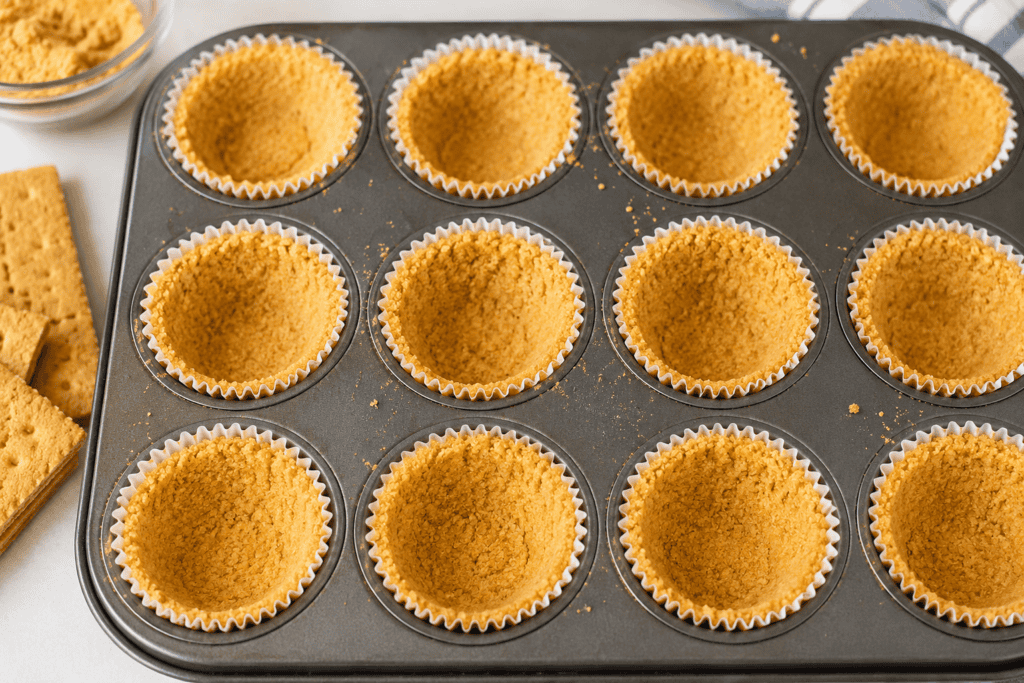 A muffin tin lined with paper cups holds mini graham cracker crust, pressed neatly in each cup, and sits on a counter beside whole graham crackers and a bowl of crumbs.