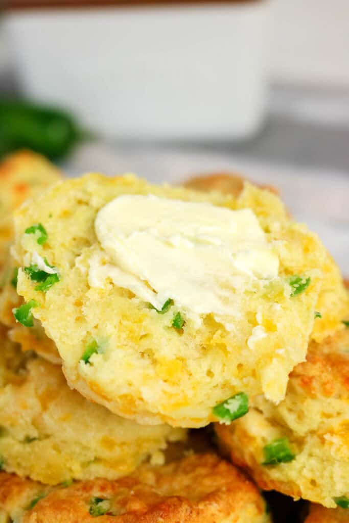 A close-up of a Jalapeno Cheddar Biscuit sliced in half, topped with a pat of melting butter. Visible green and yellow bits of jalapeños and cheese dot the biscuit, with more Jalapeno Cheddar Biscuits stacked underneath in the background.