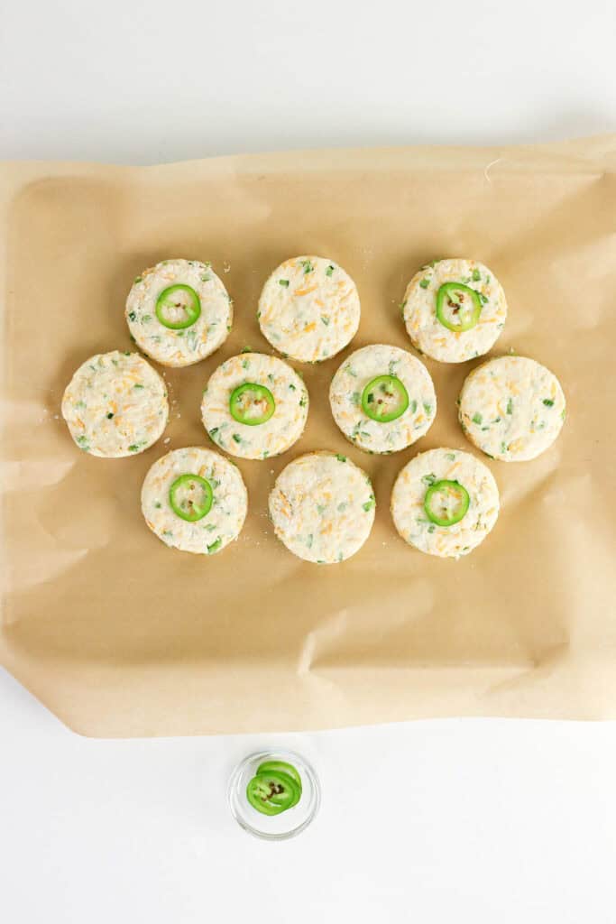 Ten round, uncooked Jalapeno Cheddar Biscuits with visible green and cheese flecks are arranged on parchment paper; three are topped with a jalapeño slice. A small bowl of additional jalapeño slices sits below them.