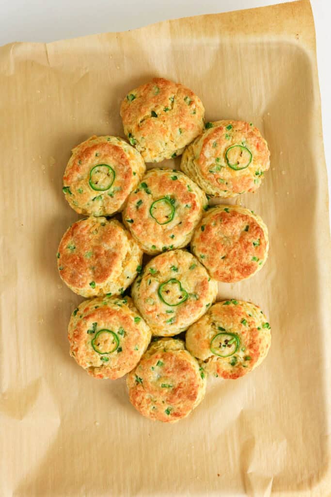 A batch of golden-brown Jalapeno Cheddar Biscuits with sliced green onions baked in, arranged closely together on a sheet of brown parchment paper.