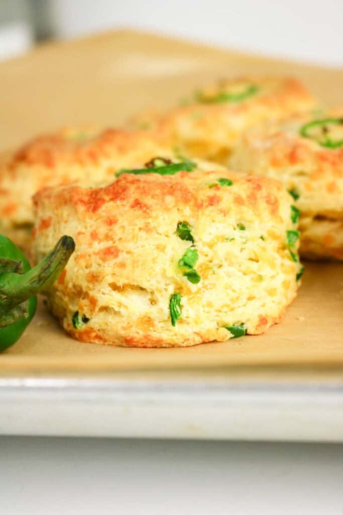 Close-up of a golden, flaky Jalapeno Cheddar Biscuit with green jalapeño pieces baked inside, resting on brown parchment paper with another biscuit and a fresh jalapeño pepper partially visible.