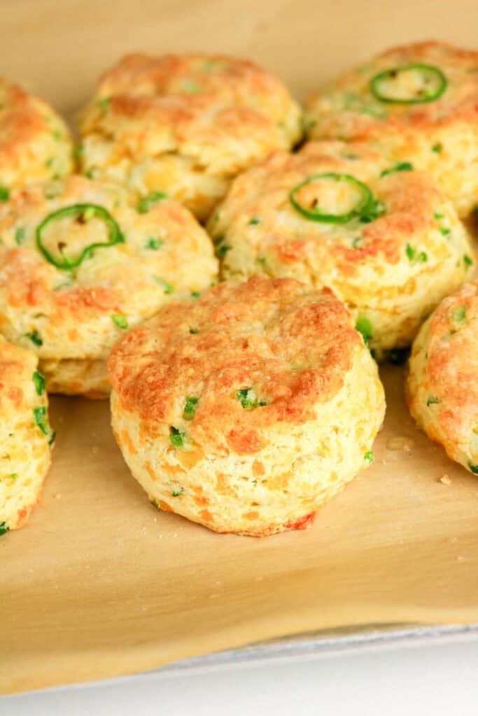 Golden brown Jalapeno Cheddar Biscuits topped with chopped green herbs and jalapeño slices, arranged closely together on a sheet of parchment paper.