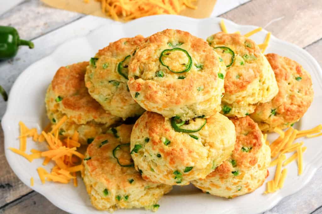 A plate of golden, freshly baked Jalapeno Cheddar Biscuits topped and filled with sliced jalapeños, surrounded by scattered shredded cheddar cheese on a rustic wooden surface.