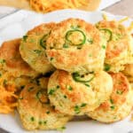 A plate of golden, freshly baked Jalapeno Cheddar Biscuits topped and filled with sliced jalapeños, surrounded by scattered shredded cheddar cheese on a rustic wooden surface.