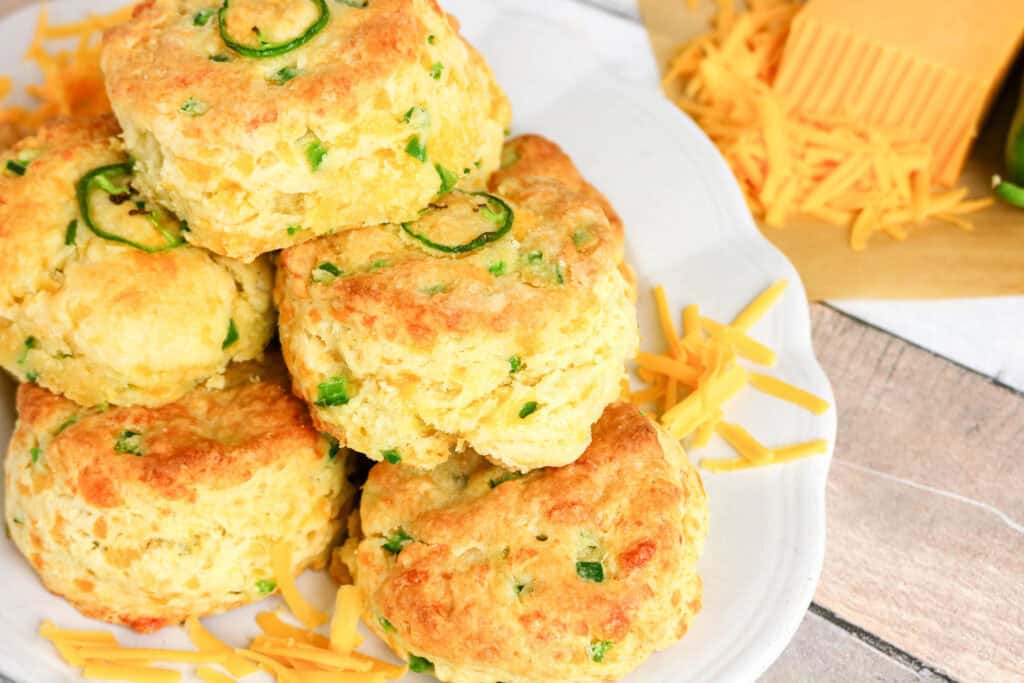 A plate of golden-brown Jalapeno Cheddar Biscuits with visible pieces of green jalapeño and shredded cheddar cheese, plus extra cheese and a block of cheddar in the background.