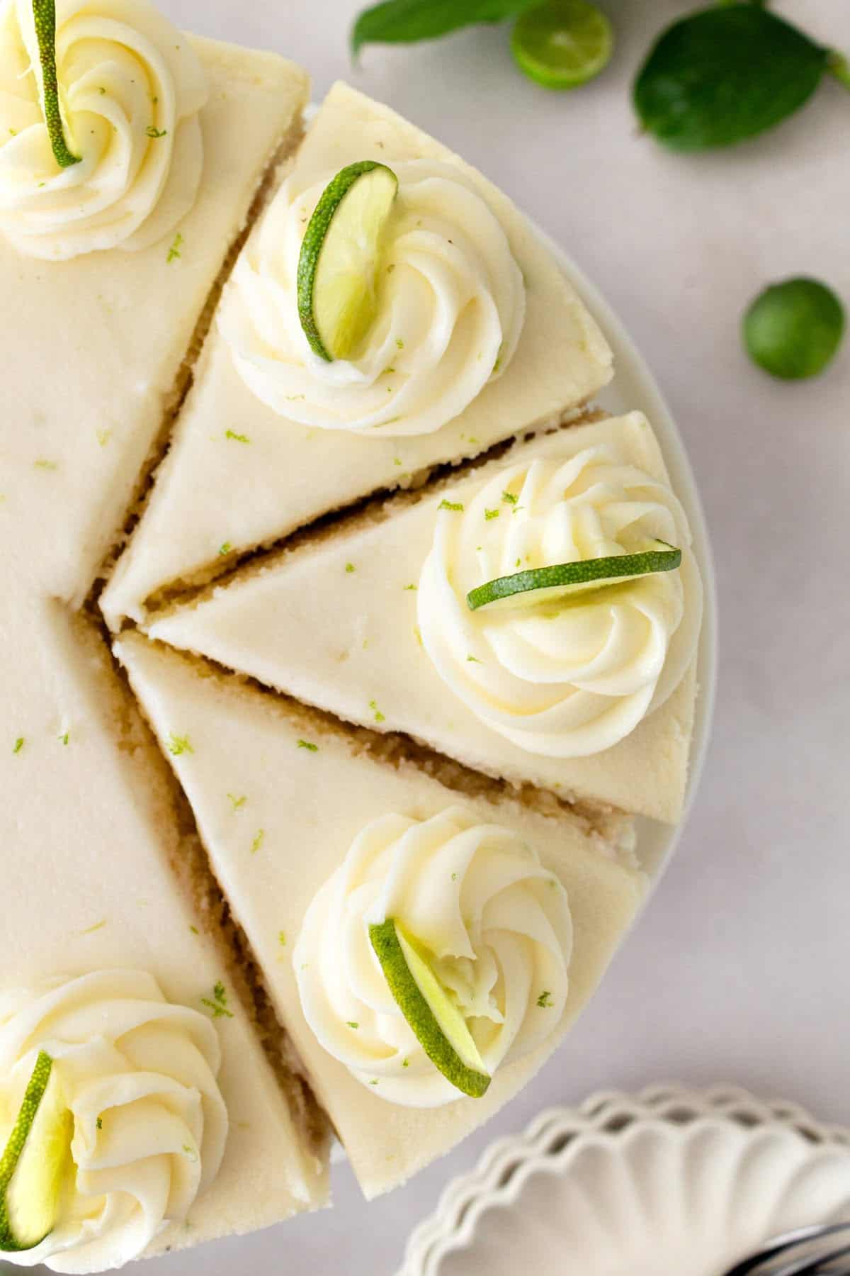 Top view of a sliced cheesecake topped with swirls of whipped cream, garnished with lime wedges and zest—an ideal choice for summer birthday cakes—displayed on a white surface with fresh green limes and leaves nearby.