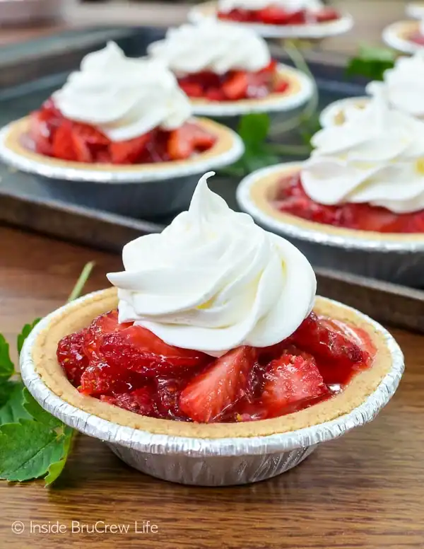 A mini strawberry pie in a foil tin with a graham cracker crust, filled with sliced strawberries and topped with a generous swirl of whipped cream, sits on a wooden table—perfect for fans of Mini Graham Cracker Crust Pie Recipes.
