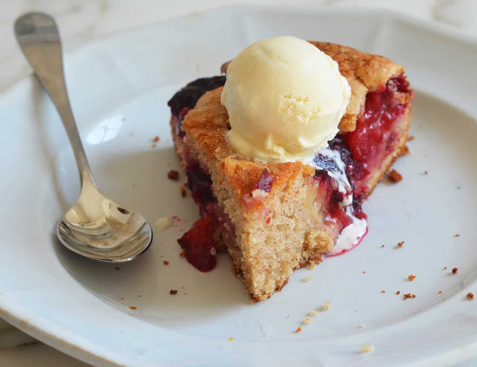 A slice of berry pie topped with a scoop of vanilla ice cream sits on a white plate next to a spoon. Some of the ice cream is melting over the warm pie, making it perfect for those who crave summer birthday cakes with a fruity twist.