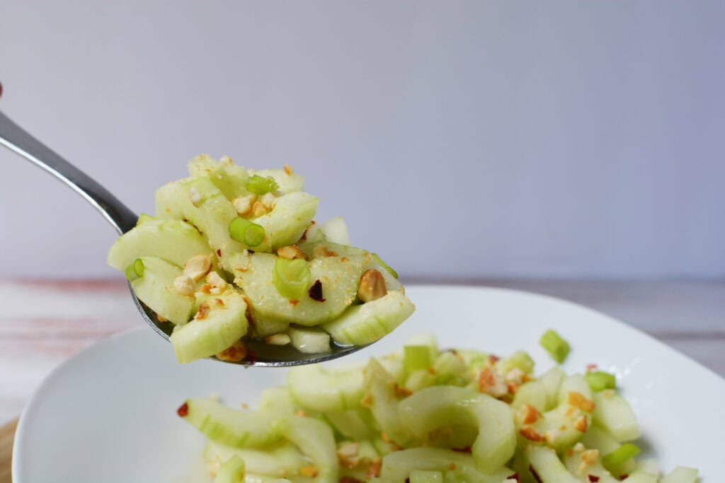 A close-up of a spoonful of Thai cucumber salad with chopped peanuts and green onions, held above a white plate filled with the same salad. The background is a light, neutral color.